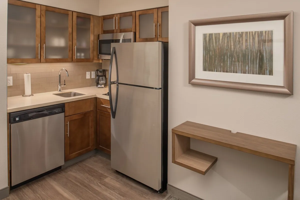 A warm, inviting kitchen interior featuring a stainless steel refrigerator, ample wooden cabinets, spacious countertops for meal prep, and cheerful morning light streaming through the window. Framed artwork and tasteful decor create a home-like environment ideal for cooking and enjoying meals together.