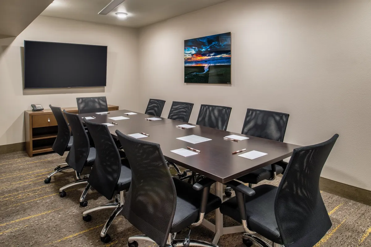 Depicts a contemporary business center with a long conference table, ergonomic chairs, a large screen TV, and tasteful wall art. The setup is ideal for meetings, brainstorming, and focused work, providing a productive yet comfortable environment.