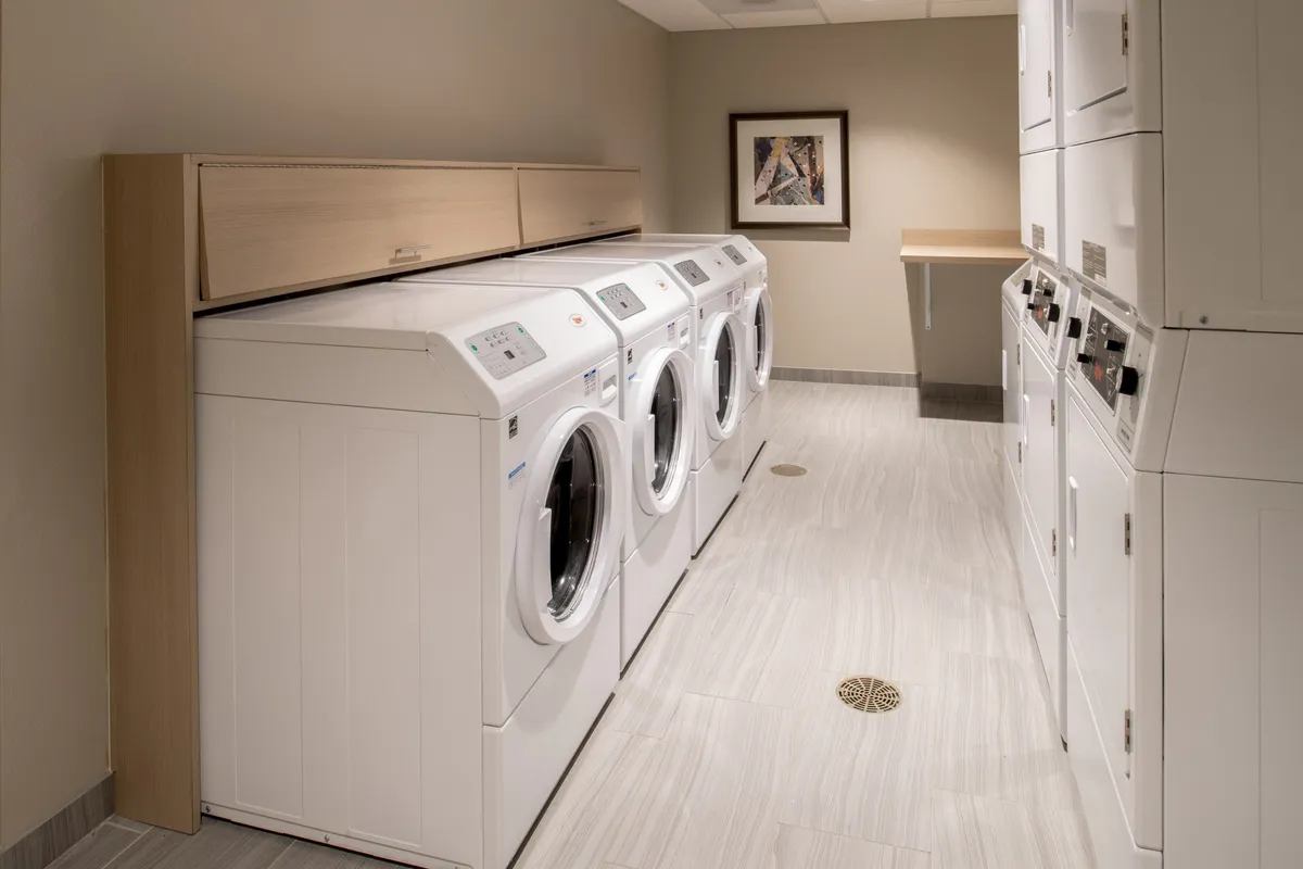 This image displays a practical and modern laundry area featuring several washing machines and dryers, complemented by a shelving unit and wall artwork. The space is well-lit and organized, reflecting a clean and efficient atmosphere that would appeal to guests needing laundry facilities during their stay.