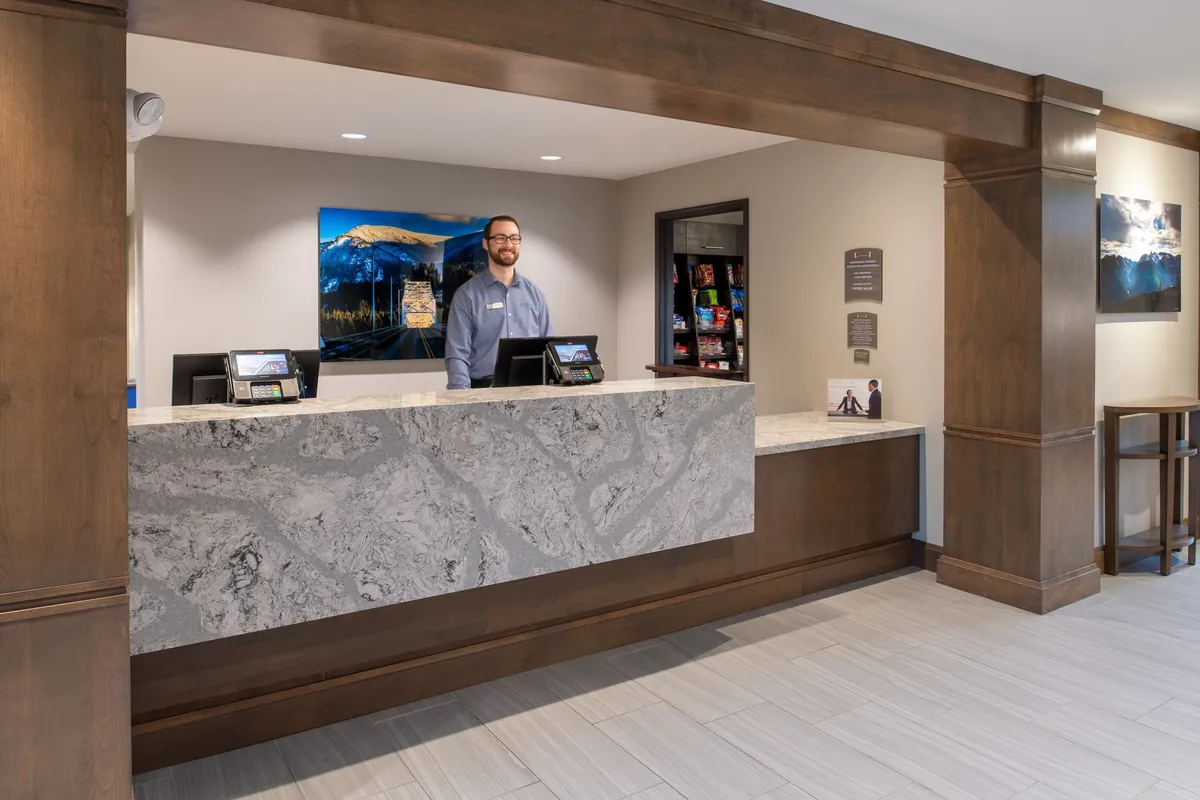 The image shows the hotel reception area, featuring a modern reception desk attended by a professional staff member. Artwork is visible on the wall behind the desk, creating a welcoming and businesslike atmosphere. The scene captures a guest being checked in or assisted, emphasizing helpful service.