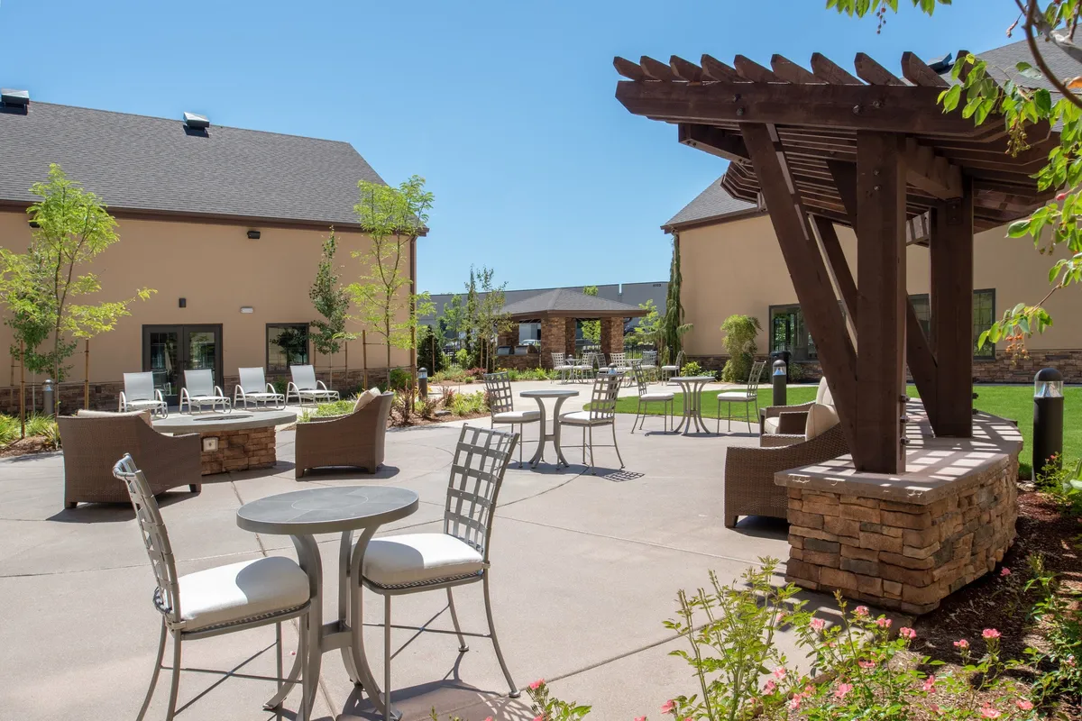 An exterior view of a pergola with outdoor seating set amid landscaped gardens, perfect for social gatherings, relaxing, or dining al fresco. The space is bathed in cheerful natural light, suggesting sunset gatherings or morning coffee outdoors.