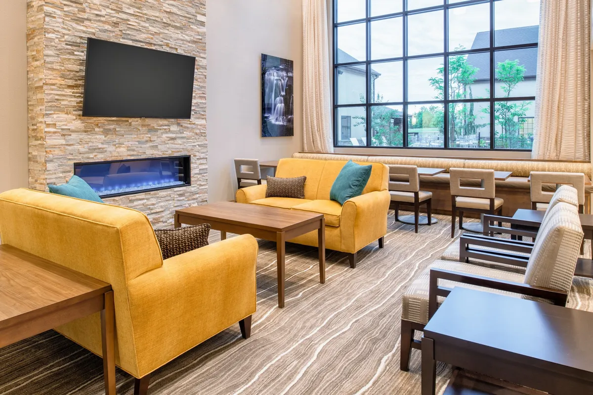 A spacious, sunlit studio lounge with plush yellow sofas arranged around modern coffee tables, large windows letting in natural light, a feature stone wall, and a flatscreen television. Tasteful artwork accents the walls, and the overall atmosphere is cozy and inviting.