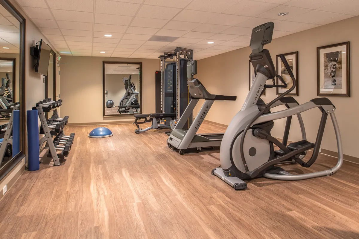 This photo captures a well-equipped fitness center featuring modern elliptical machines, a weight rack, an exercise mat, and a wall-mounted mirror. The scene conveys a motivating, active environment with guests engaging in individual workouts or stretching routines.