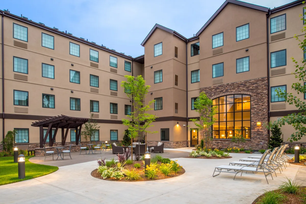 This exterior image features the stone façade of the Staybridge Hillsboro North, with large windows, a relaxed outdoor seating area, and a pergola inviting guests to enjoy the fresh air. The space suggests opportunities for families to gather outdoors and unwind after exploring nearby attractions.