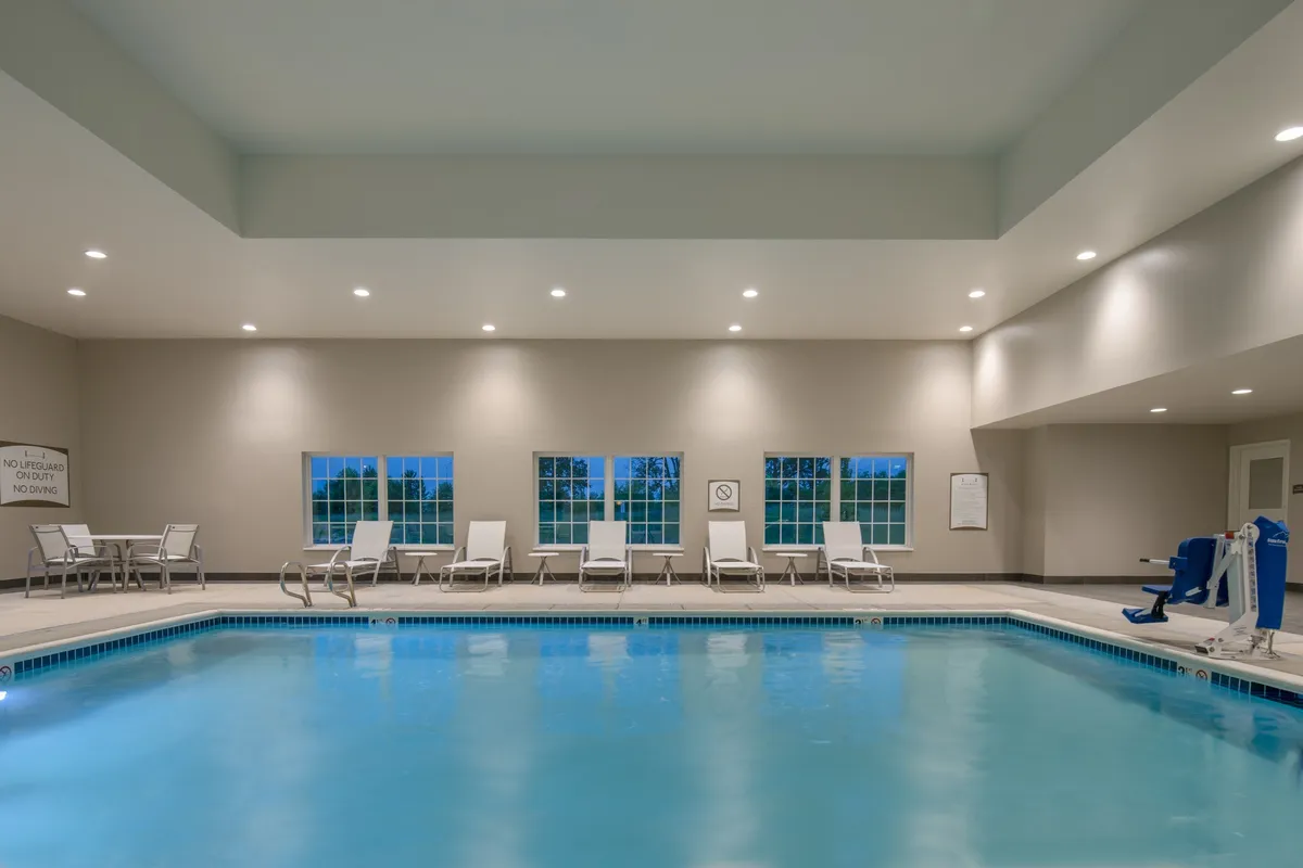 This image showcases a spacious indoor swimming pool area with crystal clear water, surrounded by lounge chairs set against large windows. The environment is tranquil, suggesting a space for both relaxation and active fun. The presence of people swimming, relaxing, and reading by the pool adds to the family-friendly, leisure-oriented atmosphere.