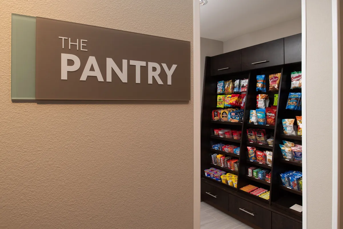 The image features 'THE PANTRY' sign above well-organized shelves filled with a variety of snacks, inviting guests to pick up treats. The scene is casual and welcoming, with people choosing snacks and socializing in a relaxed, communal atmosphere, hinting at the comforts and conveniences available to guests during their stay.